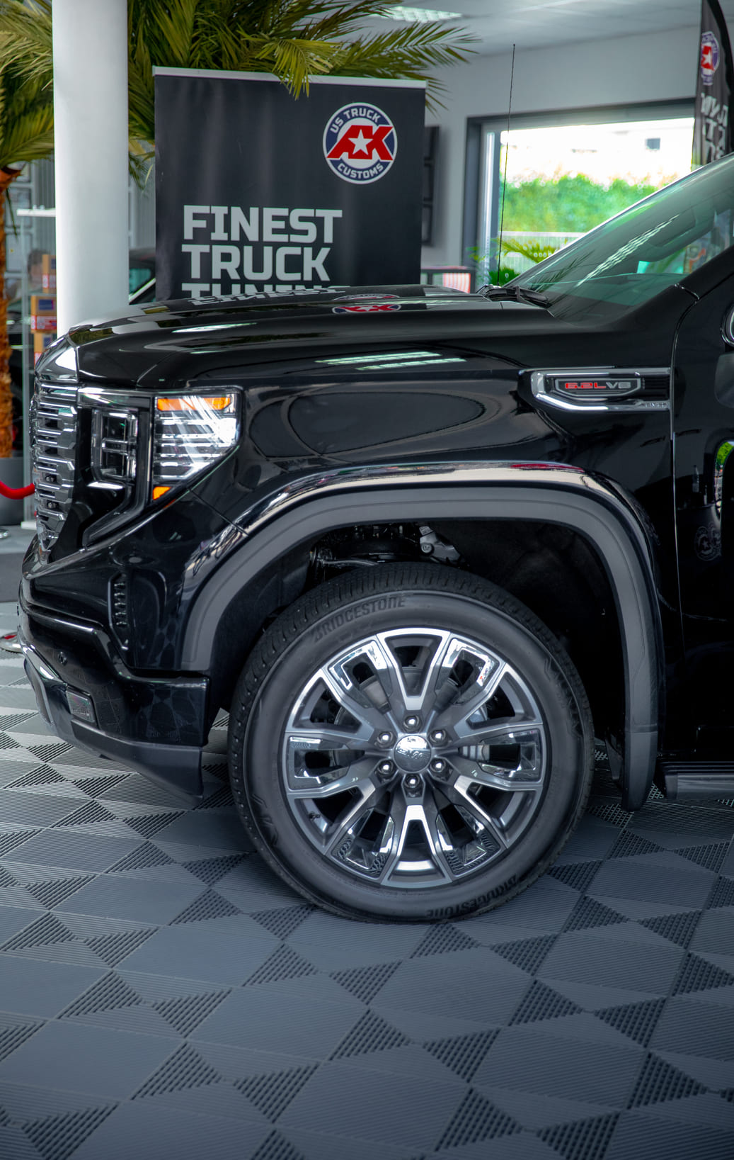 Das Bild zeigt die markante Frontpartie eines GMC Sierra 1500 Denali mit Fokus auf das vordere Rad und die Radhauspartie. Im Zentrum steht die große, polierte 22-Zoll-Denali-Felge mit Mehrspeichen-Design und Bridgestone-Bereifung. Am Kotflügel ist das „6.2L V8“-Emblem gut sichtbar – ein klares Statement für Power und Premium-Anspruch. Im Hintergrund weist ein Banner von AK Customs auf professionelles Truck-Tuning hin.